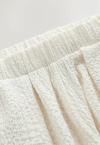 Gros plan d'un tissu texturé blanc cassé avec une ceinture élastique froncée, montrant des détails de surface froissée et plissée.
