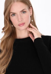 Maglione nero lavorato a maglia con scollatura a coste. Capelli lunghi e ondulati e orecchini a perla pendenti. Mano che sfiora delicatamente il viso. Pelle chiara.