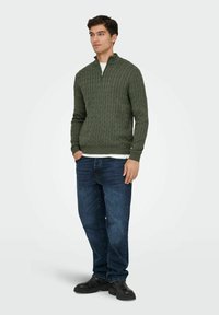 Pull en maille torsadée vert avec un col à demi-zip, assorti à un jean bleu foncé et des bottes noires. Le tissu est texturé et ajusté.