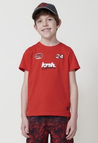 Červené bavlnené tričko s krátkymi rukávmi, na ktorom sú biele a čierne grafiky "24" a "krsh", v kombinácii s vzorovanými šortkami.