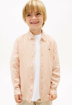 Giovane ragazzo sorridente con capelli biondi che indossa una camicia a righe arancione e bianca sopra una maglietta bianca, in piedi davanti a uno sfondo bianco uniforme.