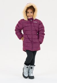 Pige iført en lila vinterjakke med hætte og pelskant, sorte leggings og grå vinterstøvler, stående med den ene hånd på hoften.