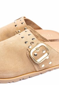 Beige suède klompen met een brede band, gouden gesp en decoratieve zilveren studs. Voorzien van een gestructureerde voetbed en een rubberen buitenzool.