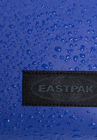 Blått tyg med en strukturerad yta, med vattendroppar och en svart logotyp som säger "EASTPAK" i botten.