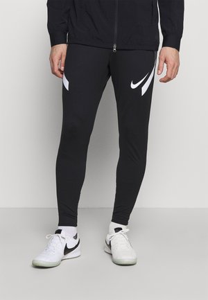 Mann trägt schwarze Nike-Leggings mit weißen Swoosh-Logos, weiße Socken und weiße Nike-Sneaker und steht auf einem einfarbigen grauen Boden.