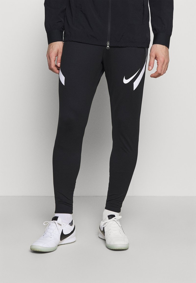 Homme portant un legging noir Nike avec des logos swoosh blancs, des chaussettes blanches et des baskets blanches Nike, debout sur un sol gris uni.