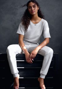 Svijetlosiva loungewear kombinacija koja se sastoji od majice opuštenog kroja i trenirki s mekim teksturom i elastičnom waistbandom. Model sjedi na tamnoj površini.