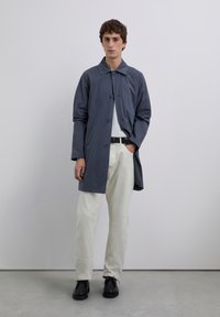 Joven de pie contra una pared gris, vistiendo un abrigo azul, camisa blanca, pantalones beige, cinturón negro y zapatos negros.