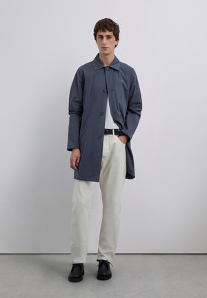 Un jeune homme se tenant contre un mur gris, portant un manteau bleu, une chemise blanche, un pantalon beige, une ceinture noire et des chaussures noires.