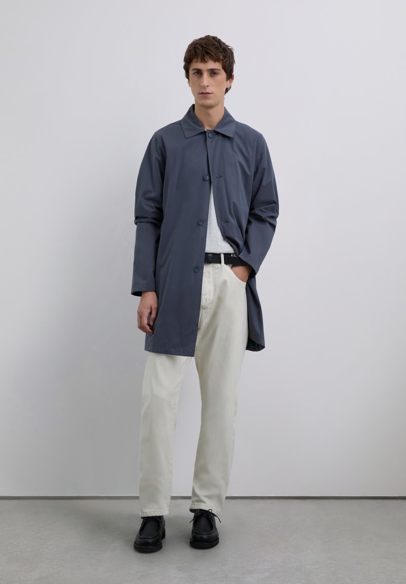 Joven de pie contra una pared gris, vistiendo un abrigo azul, camisa blanca, pantalones beige, cinturón negro y zapatos negros.