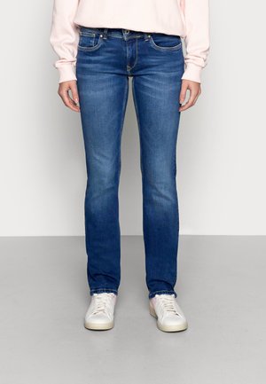 Persoon draagt blauwe slim-fit jeans, lichtroze sweatshirt en witte sneakers en staat op een grijze vloer tegen een witte muur.