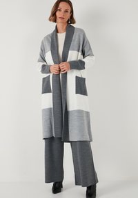 Cardigan lungo in grigio e bianco con colletto sciallato, caratterizzato da due tasche frontali e una vestibilità morbida. Realizzato in un tessuto morbido e testurizzato.