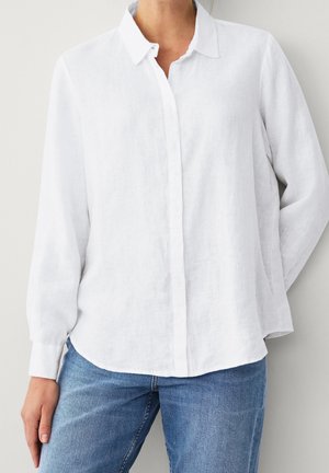 Kobieta ubrana w luźną białą koszulę z długim rękawem i guzikami, w połączeniu ze średnio niebieskimi dżinsami, stojąca na jednolitym jasnym tle.