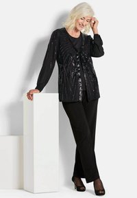 Schwarze Paillettenjacke mit langen Ärmeln und geometrischem Muster. Kombiniert mit schwarzen Hosen und High Heels. Glatte Textur, elegantes Design.