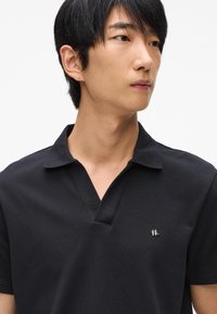 Čierna polo košeľa s textúrovanou tkaninou, krátkymi rukávmi a designom v-predu do V, s malým strieborným logom na ľavej strane hrudi.