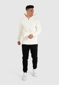 Sweat à zip blanc avec un intérieur jaune, associé à un pantalon de survêtement noir et des baskets blanches. Comprend des poches avant et une coupe décontractée.