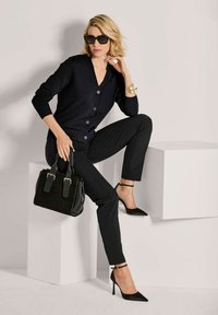 Vrouw met blond haar die een zwarte cardigan en broek draagt, zwarte hoge hakken en een zonnebril, een zwarte handtas vasthoudt en op witte blokken zit.