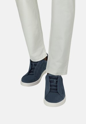 Beine in hellbeigen Hosen, die dunkelblaue Wildledersneaker mit weißen Sohlen tragen, stehen mit leicht gekreuzten Füßen auf weißem Hintergrund.