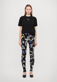 Svart t-shirt med logotyp, parat med figurnära leggings med ett färgglatt, abstrakt mönster på svart bakgrund. Svarta ballerina-skor fulländar outfiten.