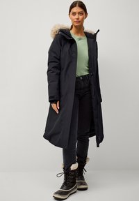 Zwarte parka met een met bont gevoerde capuchon, voorzien van een ritssluiting en zijzakken, gedragen over een groene top en donkere jeans, aangevuld met zwarte winterlaarzen.