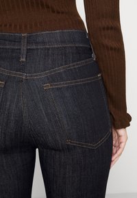 Jeans de denim oscuro con costuras naranjas, con un yugo curvado y dos bolsillos traseros, combinados con una parte superior marrón de canalé.