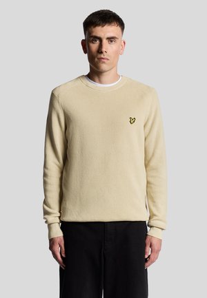 Beige ribbad långärmad tröja med rund hals och en liten gul logotyp på vänster bröst, kombinerad med svarta byxor.