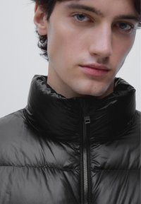 Młoda osoba o jasnej karnacji i niebieskich oczach nosi czarną błyszczącą kurtkę puchową z wysokim kołnierzem i zamkiem błyskawicznym.