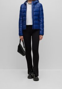 Blå dunjakke med hætte, hvid skjorte, sorte ribstrikkede leggings og sorte chunky støvler. Holder en sort håndtaske med logo-strop.