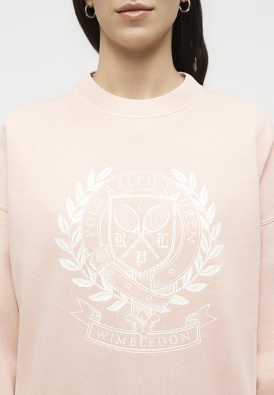 Femme portant un sweat-shirt rose clair avec un blason blanc Polo Ralph Lauren Wimbledon affichant des raquettes de tennis croisées et des feuilles de laurier.