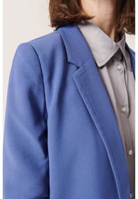 Modrá sako s měkkým texturovaným povrchem a upravenými klopami, překryté nad světle šedou košilí s knoflíky a klasickým límečkem.