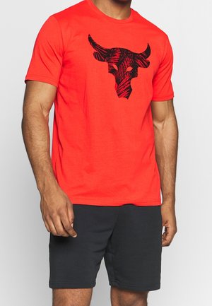Mann trägt ein knallrotes T-Shirt mit schwarzer Stierschädel-Grafik und schwarze Shorts, steht vor einem schlichten hellen Hintergrund.