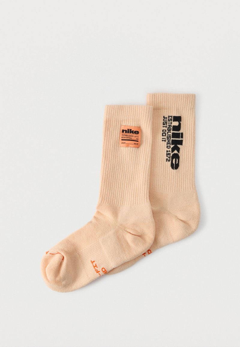 Paire de chaussettes côtelées Nike beige avec logos et texte noir et orange, posées à plat sur fond blanc.