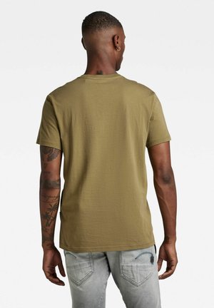 Hombre con tatuajes vistiendo una camiseta verde oliva y jeans gris claro, mostrado de espaldas sobre un fondo blanco.