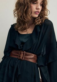 Haut noir à volants superposés en tissu texturé, porté sur une blouse en satin sombre, cintré avec une large ceinture en cuir marron ornée d'une boucle rectangulaire.