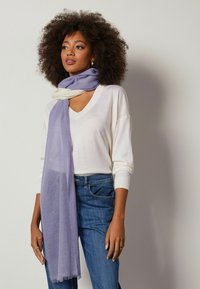 Letvægts, tofarvet tørklæde i lavendel og creme, med blød tekstur og frynser, båret løst over en hvid v-hals sweater.
