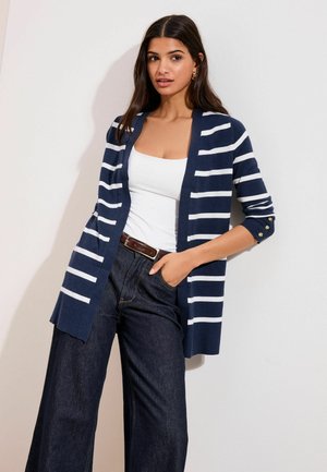 Noor naine, kellel on pikad tumedad juuksed, kannab tumesinist ja valget triibulist kardigani valge pluusi peal, tumedaid laia säärega teksapükse ning pruuni vööd, poseerides.