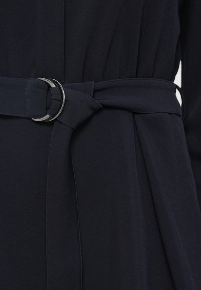 Tamsiai mėlynos audinio diržas su sidabriniu apvaliu užsegimu, surištas mazgu. Medžiaga lygi su šiek tiek blizgesiu, parodanti raukšlėtą tekstūrą.