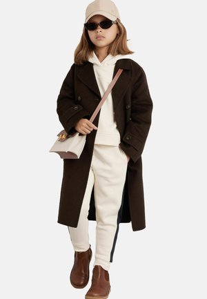 Palton din lână maronie peste un hanorac alb și pantaloni de trening crem cu dungi pe laterale. Botine maronii și o șapcă deschisă. Ochelari de soare negri.