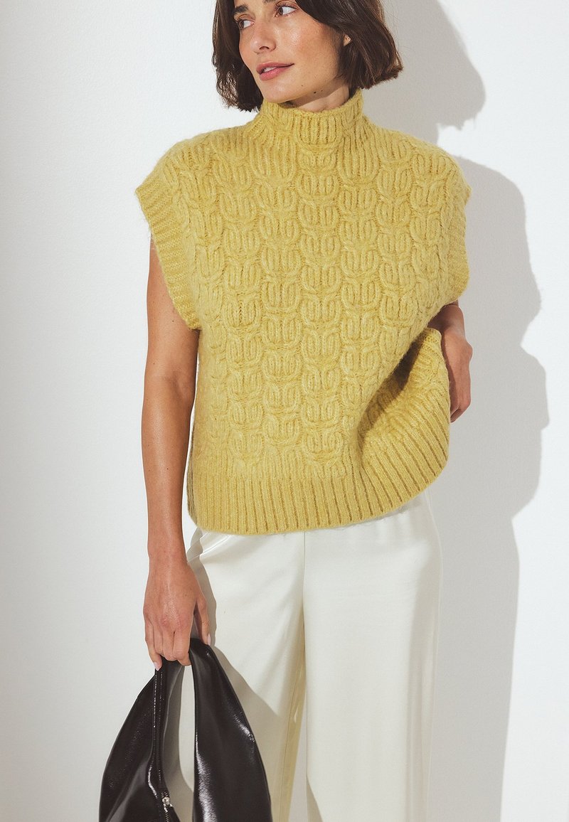 Geel gebreid sweater met een hoge halslijn en korte mouwen, met een gestructureerd patroon. Gecombineerd met witte broeken en een zwarte tas.