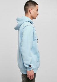 Svijetloplava hoodie s prednjim džepom, dugim rukavima i kapuljačom s uže. Ima logo sprijeda i crni detalj na manžetama.