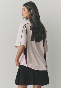 T-shirt de sport rose clair avec des accents bordeaux et un numéro 7 visible au dos, associé à une jupe plissée noire.