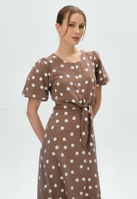 Braunes Kleid mit Pollendruck, kurzen Puffärmeln und einer gebundenen Taillenpartie. Hergestellt aus weichem Stoff mit weißen, gleichmäßig verteilten Kreisen.