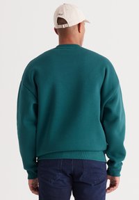 Turkusowa, oversize’owa bluza z prążkowanym dekoltem i dołem, zestawiona z ciemnymi dżinsami; noszona z jasną czapką z haftowanym napisem „OLO” z tyłu.