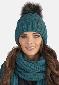 Bonnet tricoté turquoise avec un grand pompon en fausse fourrure et un patch en cuir ; porté avec une écharpe texturée assortie.