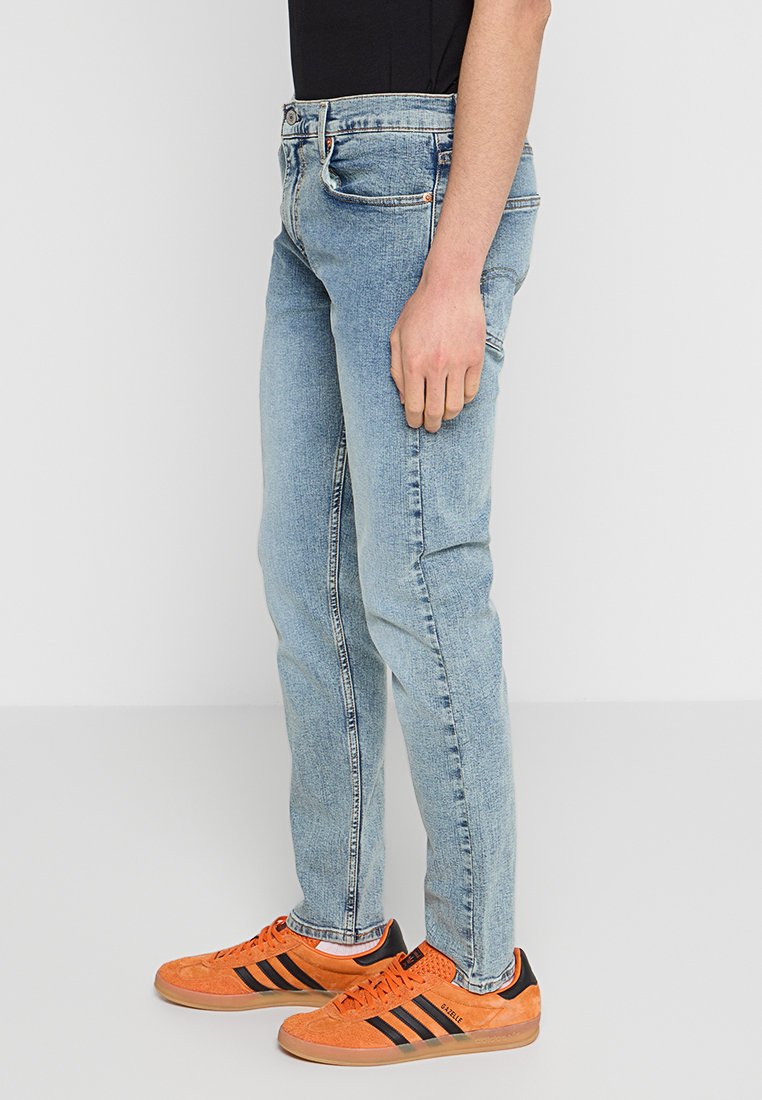 Jeans de mezclilla azul claro con corte ajustado, con un sutil desgaste y cinco bolsillos. Combinados con zapatillas de deporte naranjas y una blusa negra.