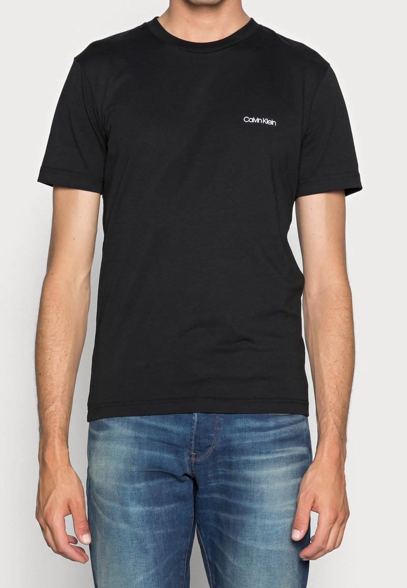 Homme portant un t-shirt à manches courtes noir Calvin Klein et un jean bleu délavé, debout avec les bras détendus le long du corps.
