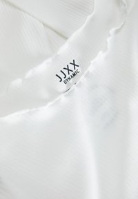 White, ribbed fabric with jagged edges; label reads "JJXX DYNAMIC" in black. Focus on texture and material, featuring a clean design.