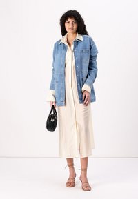 Denimjakke med lys blå vask, lange ermer, brystlomme, lagdelt over en beige midi-kjole, parret med sandaler med stropper og en liten svart veske.