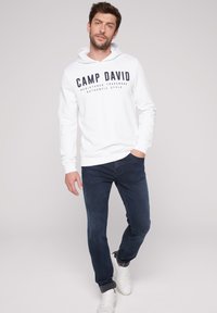 Witte hoodie met 'CAMP DAVID' in donkerblauw gedrukt op de voorkant, gecombineerd met donkerblauwe jeans en witte sneakers. Casual pasvorm en ontwerp.