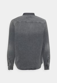 Camisa de mezclilla gris con cuello abotonado, mangas largas y un dobladillo ligeramente curvado. Presenta un desgaste y una etiqueta en la parte posterior.